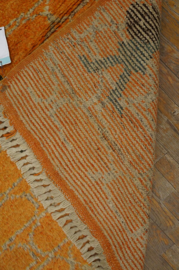High pile orange rug with gray pattern is folded to show the multicolored striped woven underside and cream fringes.