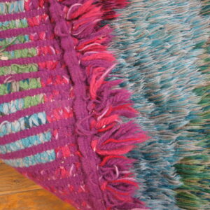 The rug edge displays magenta stripes and fringe next to long blue and green shaggy texture on wood.
