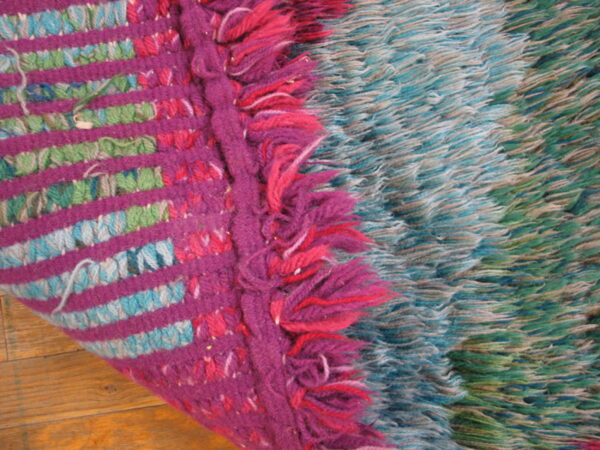 The rug edge displays magenta stripes and fringe next to long blue and green shaggy texture on wood.