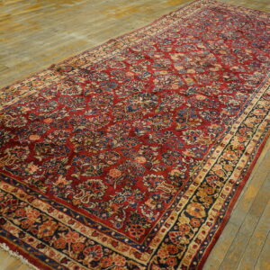 Red rug with a repeating botanical pattern in blue and beige lies on worn hardwood floors.