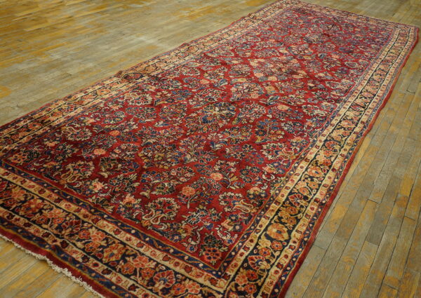 Red rug with a repeating botanical pattern in blue and beige lies on worn hardwood floors.
