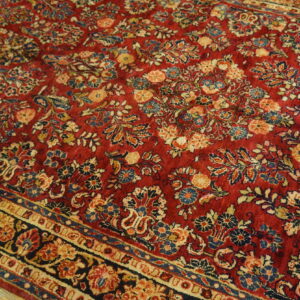 Textured red rug with intricate gold and blue botanical motifs and a decorative border shown on wood flooring.