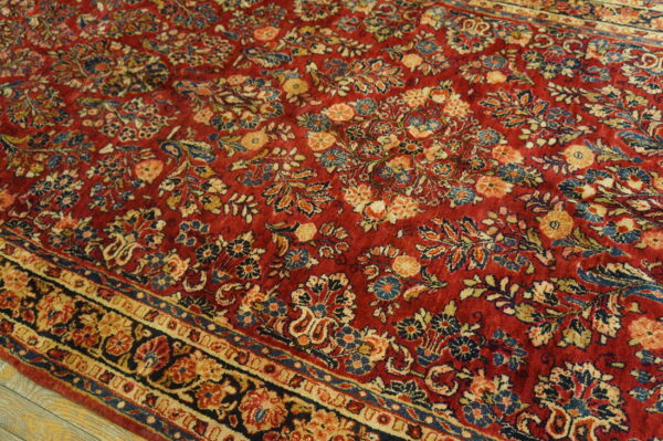 Textured red rug with intricate gold and blue botanical motifs and a decorative border shown on wood flooring.