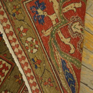A low-pile red, olive, and blue patterned rug with fringes is rolled on rustic wood planks.
