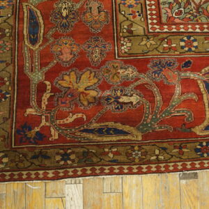 Close-up of a red rug corner with intricate blue, gold, and green floral patterns on distressed wood flooring.