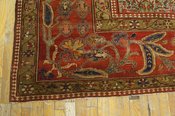 Close-up of a red rug corner with intricate blue, gold, and green floral patterns on distressed wood flooring.