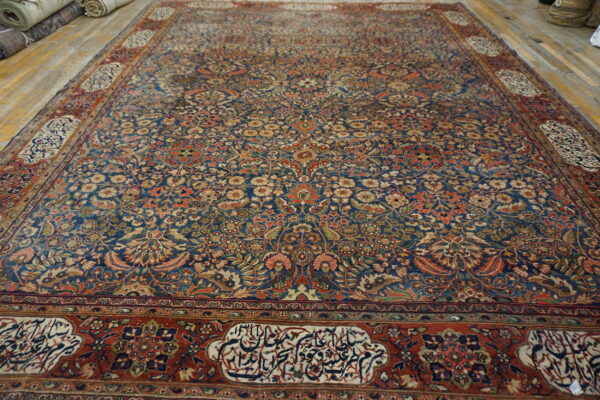 Large blue rug with a dense traditional floral pattern and rust borders featuring script cartouches on a wood floor.