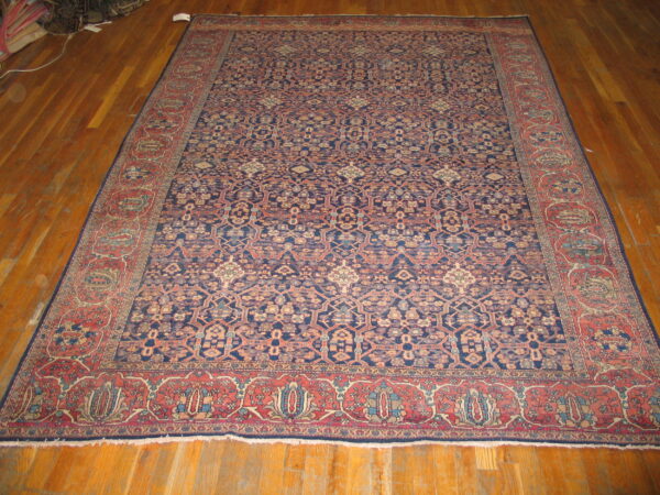 Dark blue patterned rug with repeating motifs and wide red borders rests on brown hardwood floors.