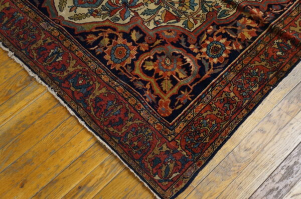 Low-pile rug corner with intricate navy, red, and gold floral patterns rests on distressed hardwood flooring.