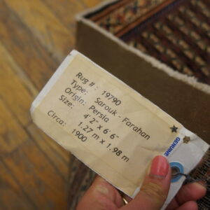Close-up of a hand holding a descriptive paper tag attached to the edge of a patterned rug on wood flooring.