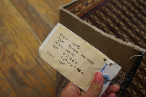 Close-up of a hand holding a descriptive paper tag attached to the edge of a patterned rug on wood flooring.