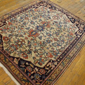 A square rug with a beige floral field and dark navy border rests on worn wood flooring.