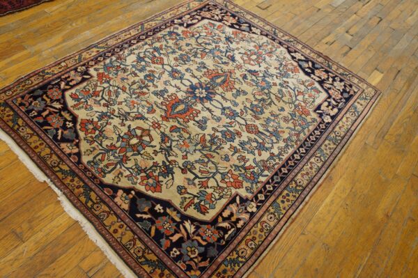 A square rug with a beige floral field and dark navy border rests on worn wood flooring.