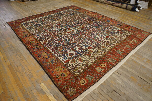Low-pile rug with an ivory field, botanical patterns, and wide reddish-orange borders on wood planks.