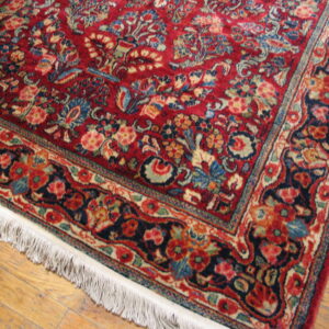 Deep red rug with complex blue and cream floral motifs, bordered by white fringe on wood floors.