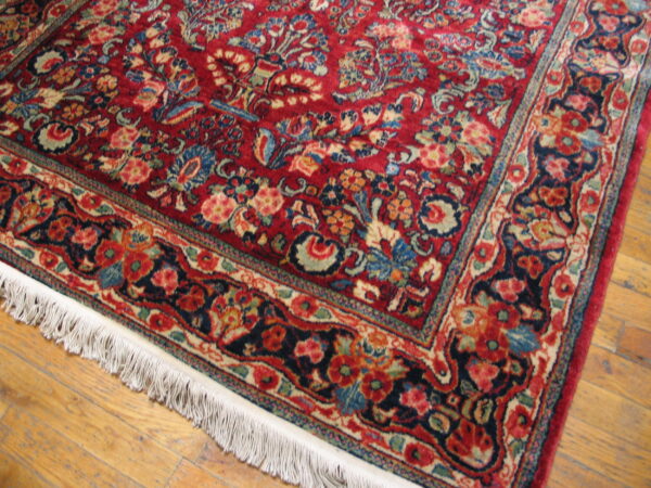 Deep red rug with complex blue and cream floral motifs, bordered by white fringe on wood floors.