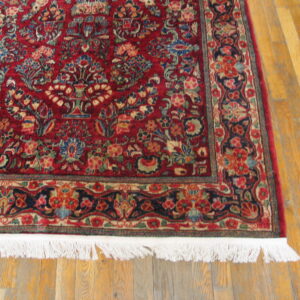 Close up of a red and navy floral rug with white fringe lying on light wood planks.