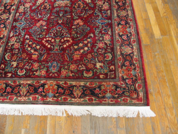 Close up of a red and navy floral rug with white fringe lying on light wood planks.