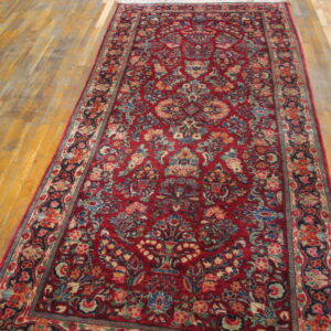A red runner rug with a dense multicolor floral pattern is laid on light brown wooden plank flooring.