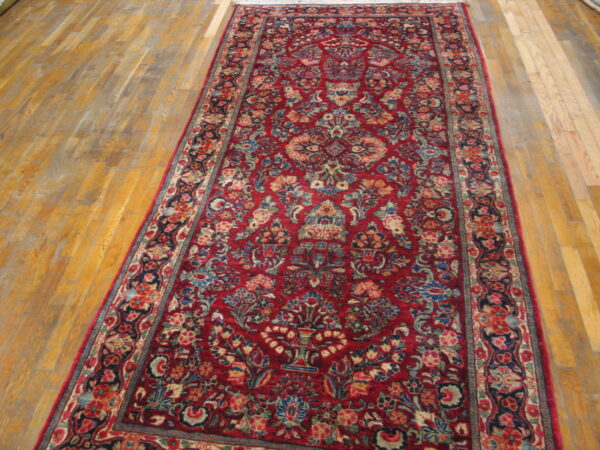 A red runner rug with a dense multicolor floral pattern is laid on light brown wooden plank flooring.