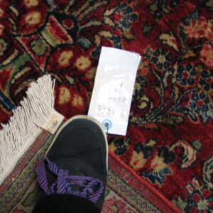A traditional patterned, high-pile red rug displays its fringe, tag, and a black sneaker with purple laces.