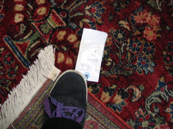 A traditional patterned, high-pile red rug displays its fringe, tag, and a black sneaker with purple laces.