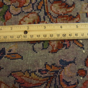 Close-up of a dense, short-pile rug pattern in gray, red, and blue, measured by a visible wooden ruler.