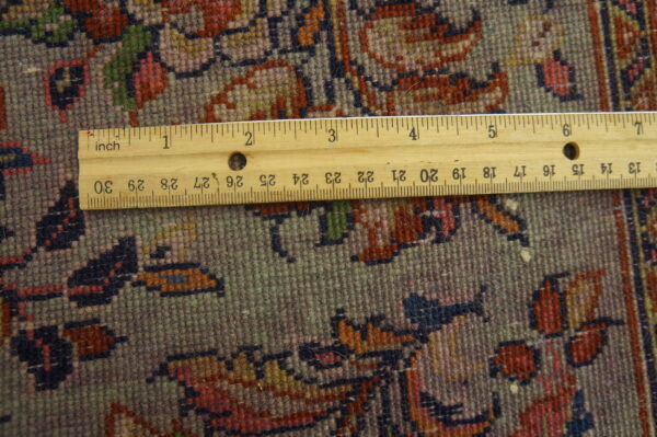 Close-up of a dense, short-pile rug pattern in gray, red, and blue, measured by a visible wooden ruler.