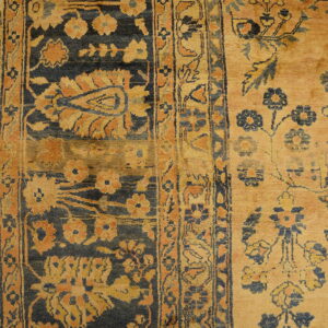 Golden tan rug featuring dark blue and rust floral motifs, detailed borders, resting on wood plank flooring.