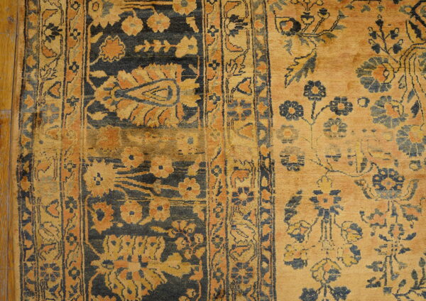 Golden tan rug featuring dark blue and rust floral motifs, detailed borders, resting on wood plank flooring.