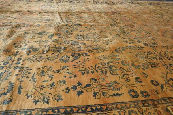 Angled view of a low-pile saffron rug featuring a complex dark blue and rust botanical pattern.