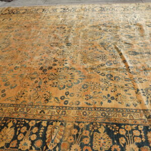 Large orange and blue low-pile rug with a dense floral pattern and wide dark border on concrete flooring.