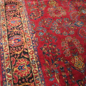 Ornate red rug with a vase and floral pattern, featuring black, blue, and gold borders, on wood planks.