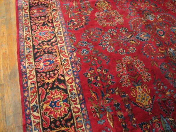 Ornate red rug with a vase and floral pattern, featuring black, blue, and gold borders, on wood planks.