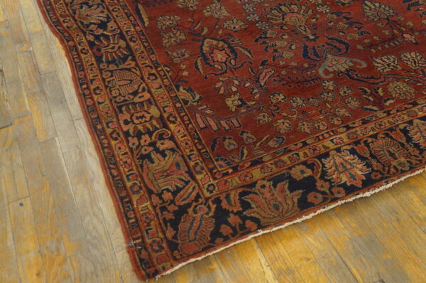 The corner of a low-pile rust red rug with detailed dark blue and gold motifs rests on light wood flooring.