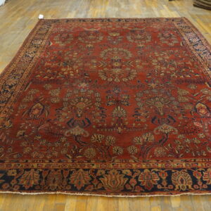 Large rust red rug with detailed blue and gold botanical patterns displayed on yellow hardwood flooring.