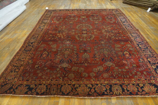 Large rust red rug with detailed blue and gold botanical patterns displayed on yellow hardwood flooring.