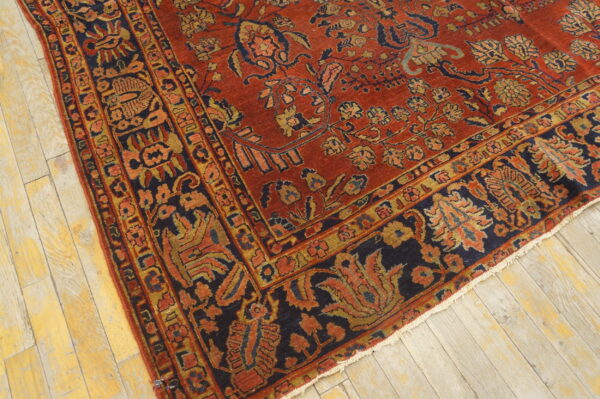Low-pile rug in deep red, navy blue, and gold floral patterns rests on distressed light wooden plank flooring.