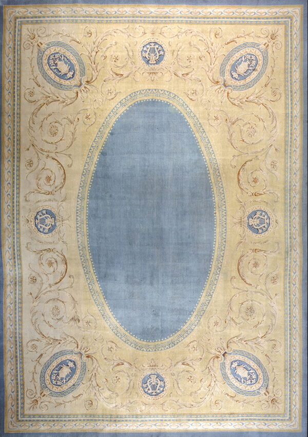 Pale yellow rug with a solid blue central oval and elaborate scrollwork featuring blue classical medallions.