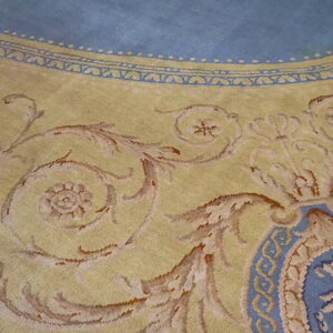 Ornate rug detail in yellow and light blue, showing scrolling foliate motifs and an intricate border design.