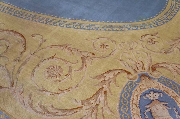 Ornate rug detail in yellow and light blue, showing scrolling foliate motifs and an intricate border design.