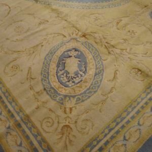 Close-up of a pale yellow rug featuring blue and tan baroque scrolling patterns and a central urn medallion on wood flooring.