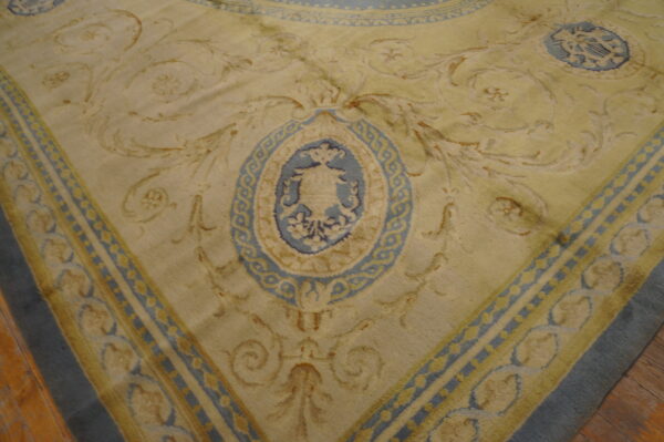 Close-up of a pale yellow rug featuring blue and tan baroque scrolling patterns and a central urn medallion on wood flooring.