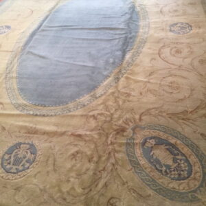 Ornate area rug featuring a blue central oval and scrollwork on a gold field laid over a wood floor.