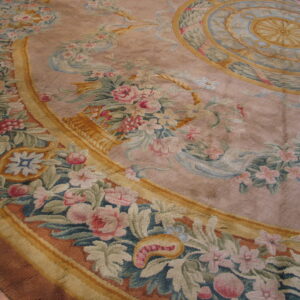 Detailed floral and ribbon medallion rug in faded pink, green, and gold colors resting on a wood floor.