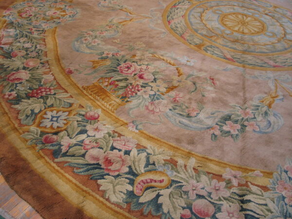 Detailed floral and ribbon medallion rug in faded pink, green, and gold colors resting on a wood floor.