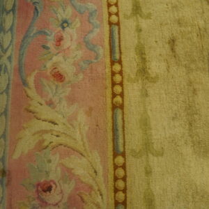 Close view of a pink floral rug border transitioning to a muted gold field on yellow wood planks.