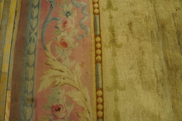 Close view of a pink floral rug border transitioning to a muted gold field on yellow wood planks.