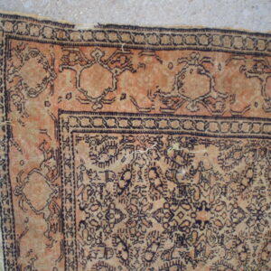 Distressed apricot and tan patterned rug corner with dark outlining and light fringe on concrete.