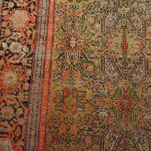 Intricate traditional rug pattern in mustard, black, and rust colors against distressed gray wood planks.